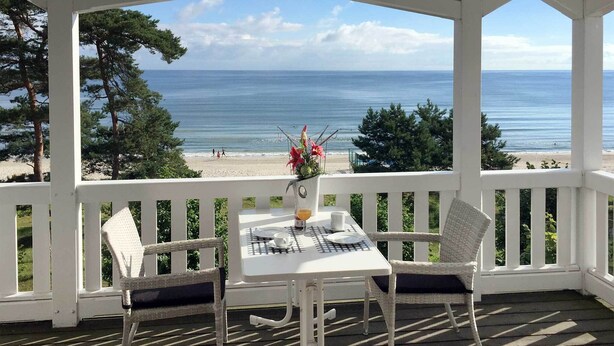Un espace extérieur abrité, avec une table et des chaises, offrant une vue sur une plage et l’océan.