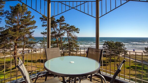 Ein überdachter Außenbereich mit Glastisch und Stühlen, mit Blick auf einen Strand und das Meer.