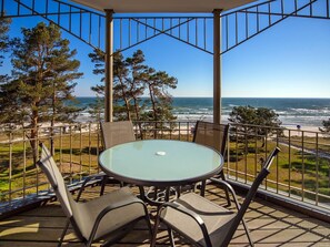 Zona d'estar exterior coberta amb taula i cadires de vidre, amb vista a una platja i l'oceà.