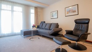 Ein modernes Wohnzimmer mit einem grauen Ecksofa, einem schwarzen Ledersessel und einem kleinen runden Tisch mit Blumen.