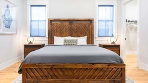 A bedroom with a wooden bed, two bedside tables, a nightstand, a patterned pillow, and a framed picture on the wall.