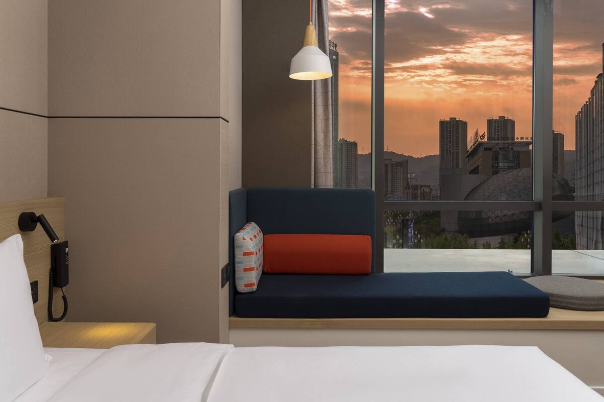 Habitación de hotel con cama, teléfono junto a la cama y un asiento junto a la ventana con vistas al horizonte urbano al atardecer.