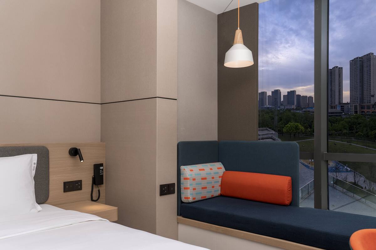 Habitación de hotel con cama, teléfono junto a ella, zona de estar con cojín y vistas a un paisaje urbano a través de amplias ventanas.