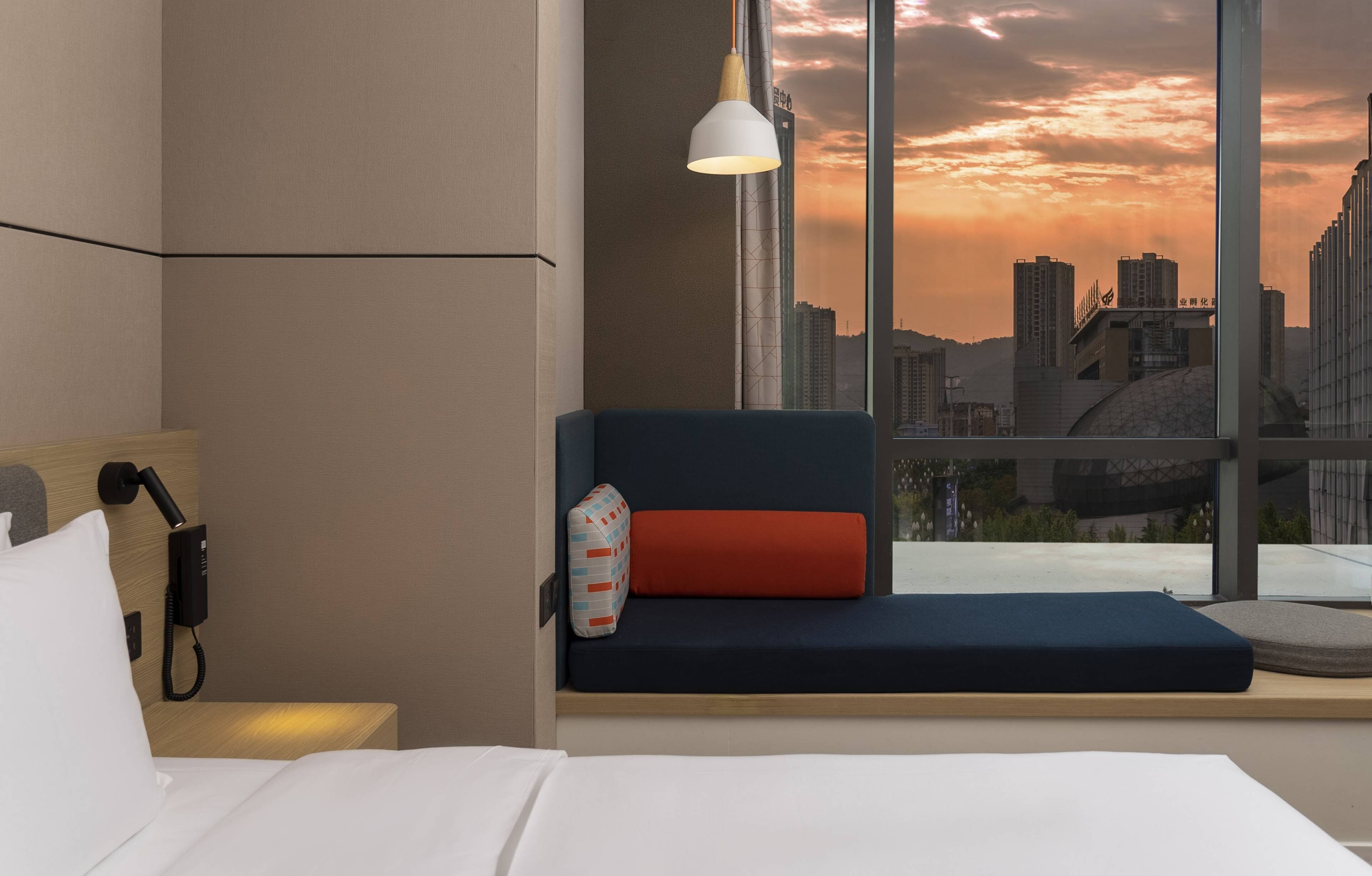 A hotel room with a bed, a bedside telephone, a window seat with a cushion, and a view of a cityscape at sunset.