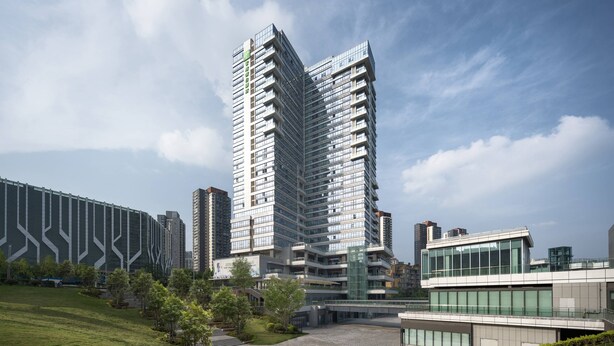 Ein modernes Hochhaus mit mehreren Etagen und Balkonen, umgeben von Grünflächen und weiteren Gebäuden.