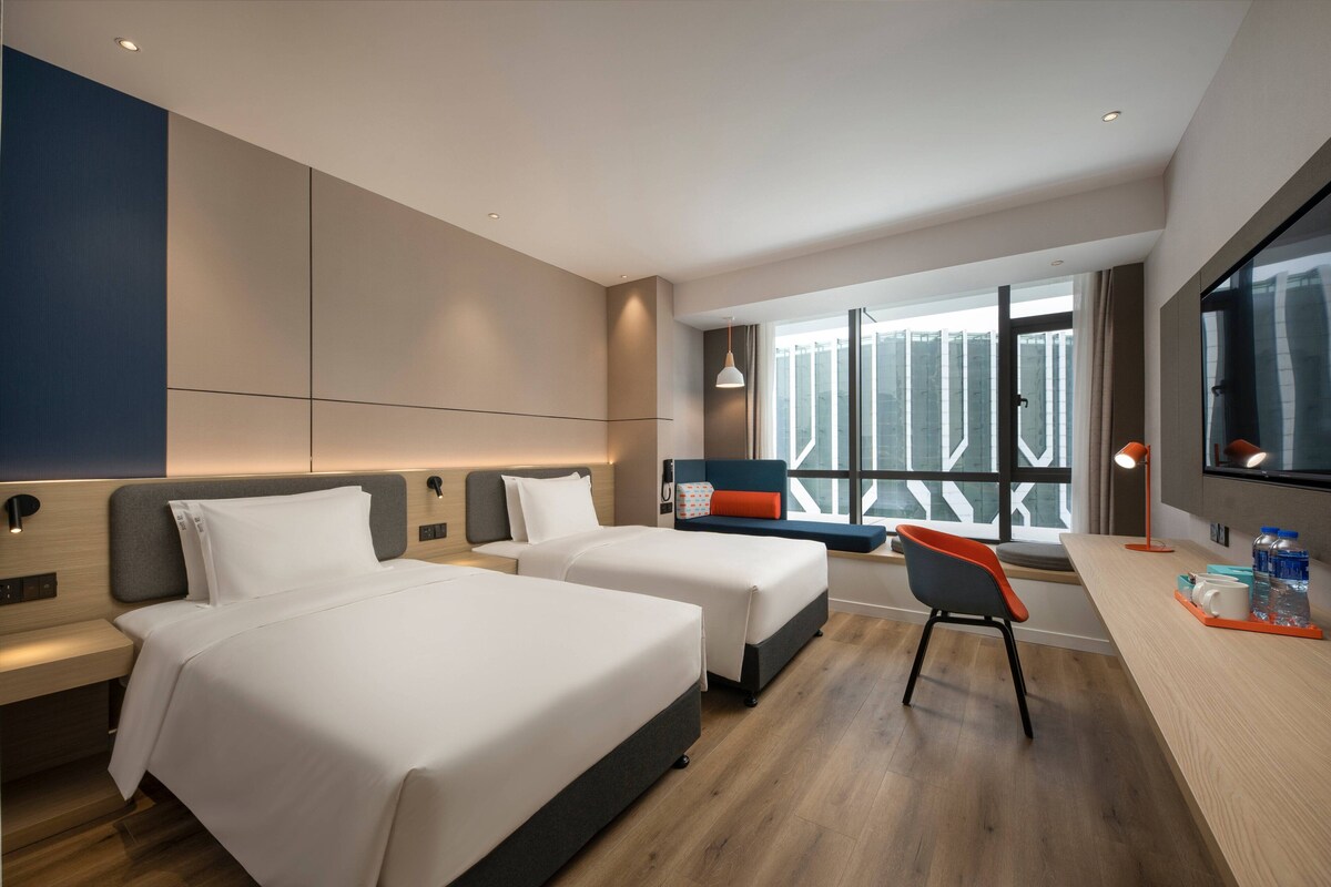 Een moderne hotelkamer met twee bedden, een bureau, een stoel en een groot raam met uitzicht op gebouwen.