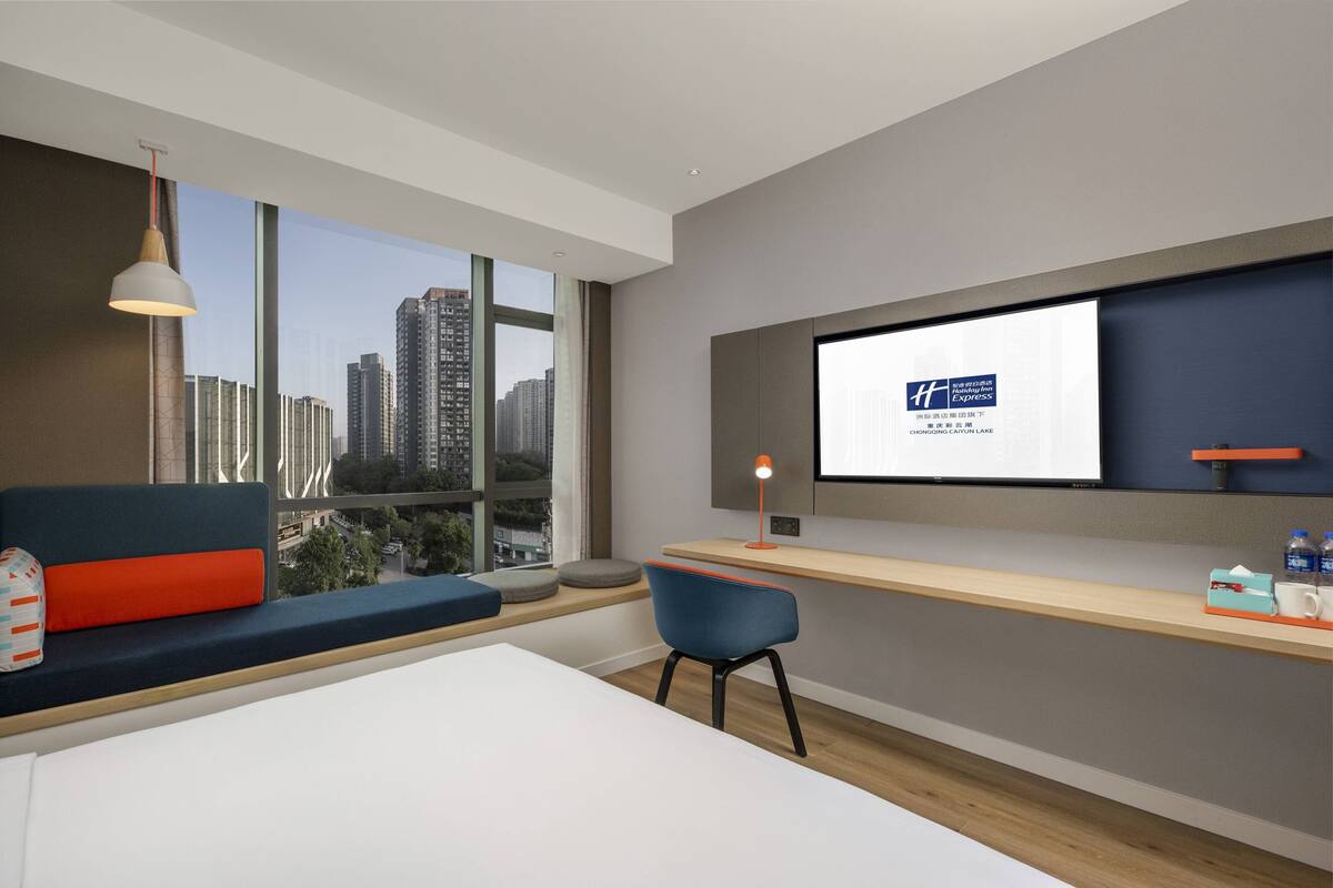 Habitación de hotel moderna con un amplio ventanal que ofrece vistas a la ciudad, un área de estar cómoda, un escritorio con ordenador y un televisor de pared.