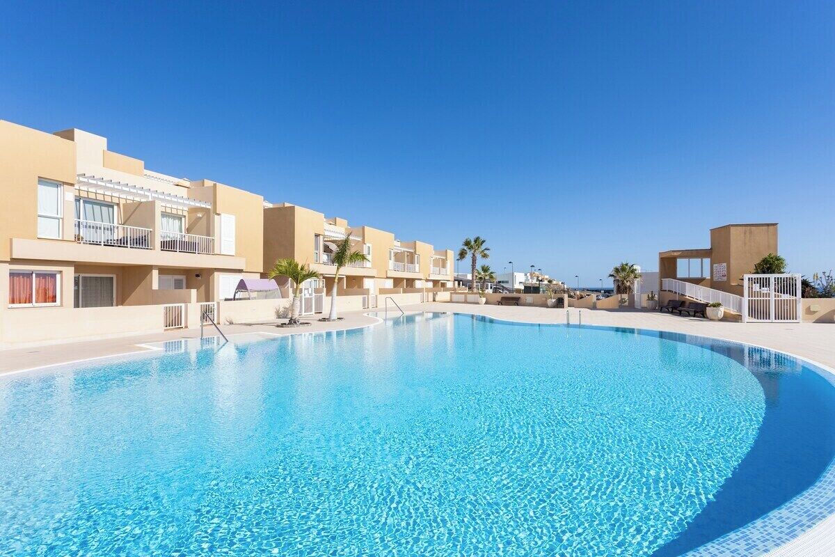 Une piscine entourée de bâtiments modernes dotés de balcons, sous un ciel dégagé, avec des palmiers.
