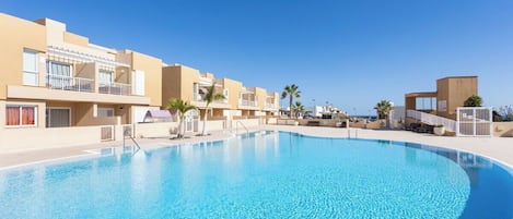 A swimming pool surrounded by modern buildings with balconies, clear skies, and palm trees.