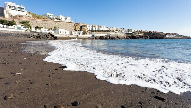A beach with dark sand, waves, and buildings in the background. - Home2Book Stunning Sea Views El Porís (Porís de Abona)