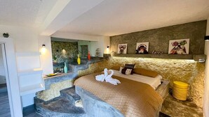 Un dormitorio con pared de piedra, una cama con manta color beige y una escalera que lleva a un balcón.