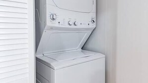 A white front-loading washer and dryer unit with control panel, located in a corner of a room with wooden flooring and white walls.