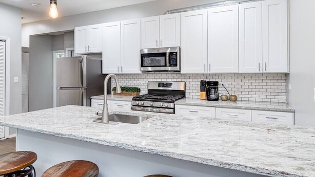 A modern kitchen with white cabinets, a stainless steel refrigerator, a microwave, a stove, and a sink. There are wooden stools at the island.
