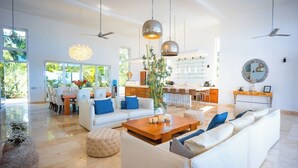Un salón amplio con un sofá grande, una mesa de centro y una zona de comedor con sillas. Se observa una cocina al fondo con una barra.