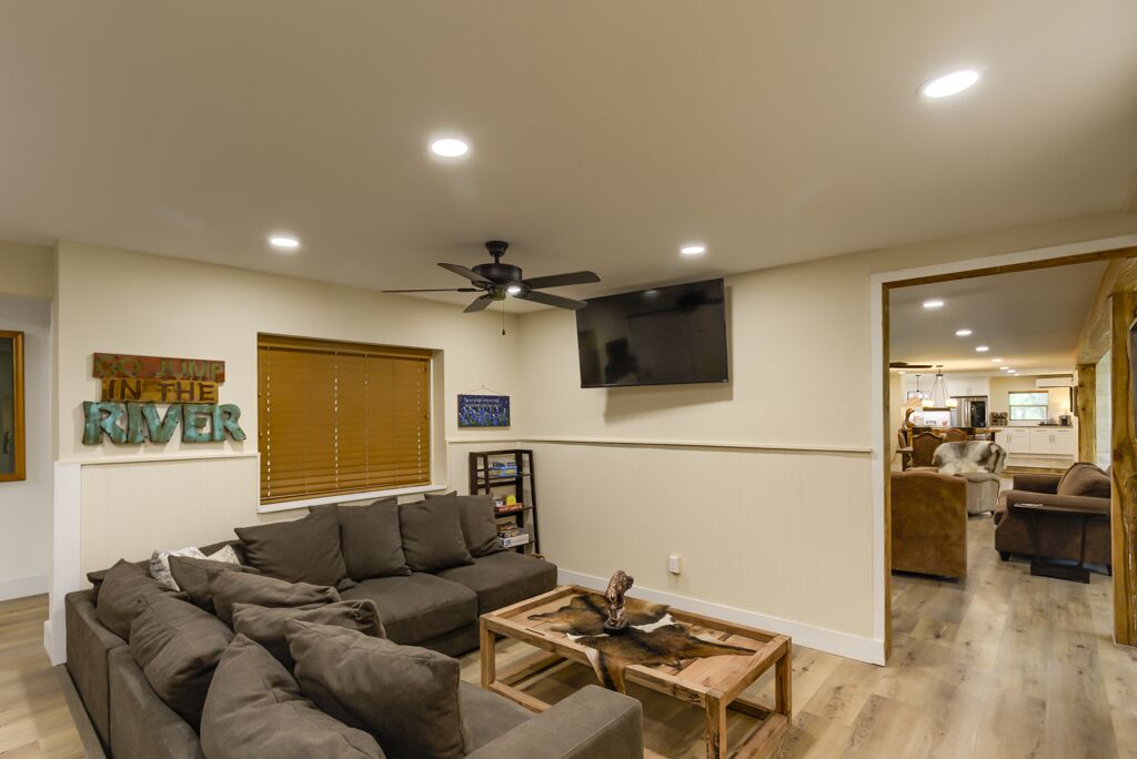 A modern living room with a sectional sofa, a coffee table, and a ceiling fan. There is a television mounted on the wall and a window with blinds.