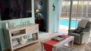 A living room with a TV, white furniture, a coffee table, a sofa, and a view of a swimming pool.