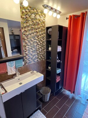 Un baño con salpicadero de azulejos, un lavamanos, un espejo, una cortina de ducha y un estante con toallas.