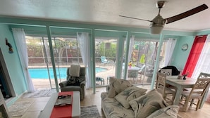 A living room with a sofa, a coffee table, and a dining area with a table and chairs. There is a ceiling fan and a view of a swimming pool through glass doors.
