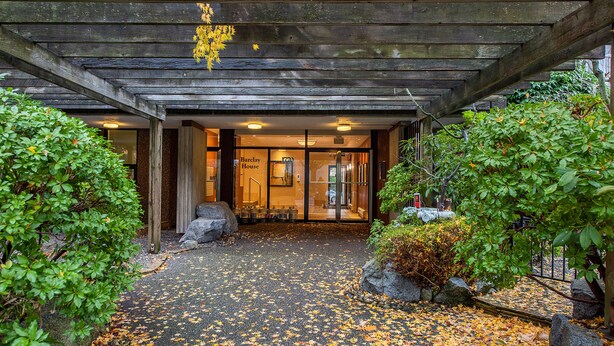 A wooden entrance with a covered walkway, surrounded by greenery and a path covered with fallen leaves.