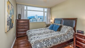 A bedroom with a large bed, a dresser, a nightstand, a lamp, and a view of buildings through the window.