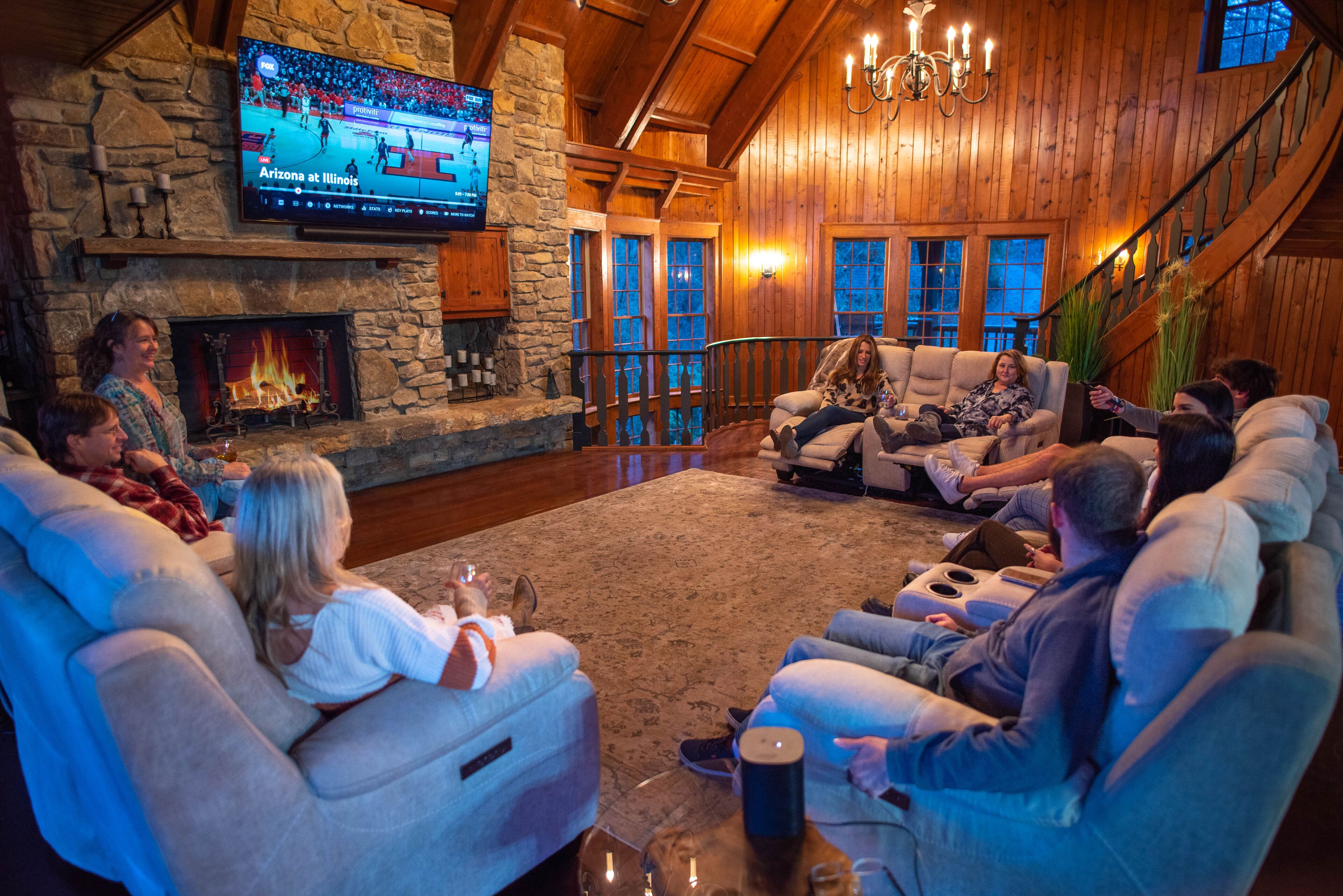 Ein gemütliches Wohnzimmer mit Kamin aus Stein, bequemen Sitzmöglichkeiten und einem großen Fernseher, der ein Sportspiel zeigt.