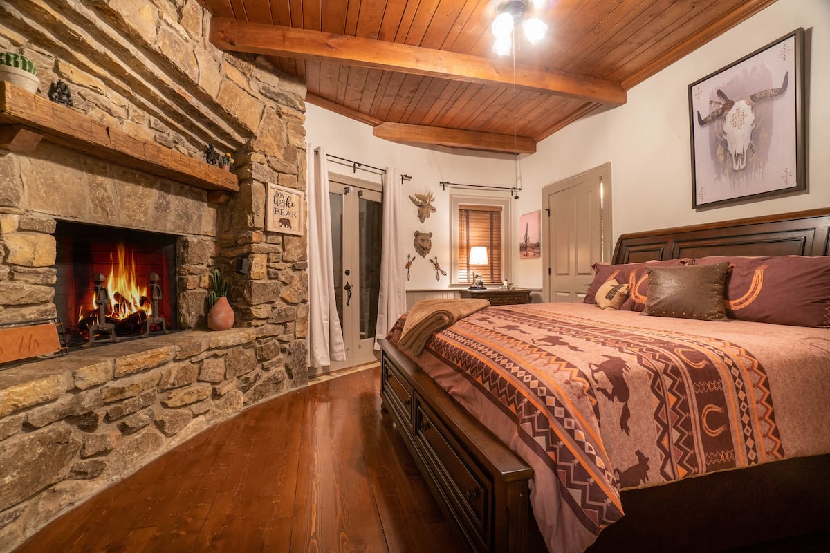 A bedroom with a stone fireplace, wooden ceiling beams, a large bed with patterned bedding, and a wall-mounted bull skull artwork.