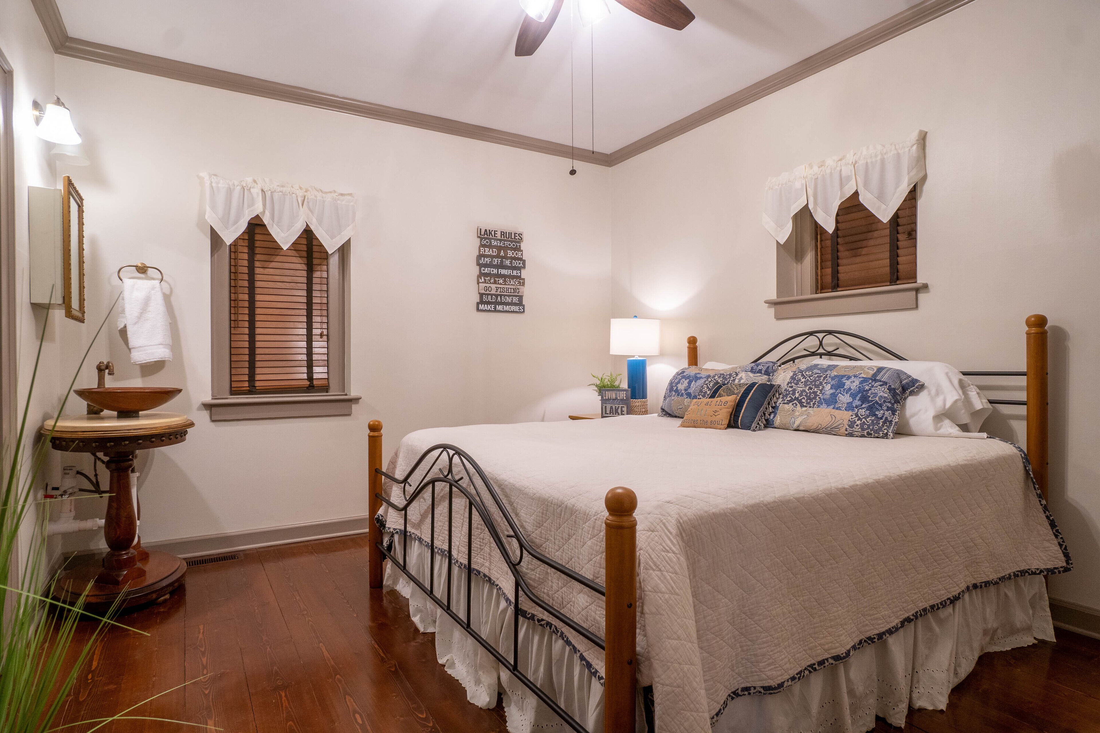 A bedroom with a wooden bed, two windows with white valances, a bedside table with a lamp, and a wall-mounted sign.