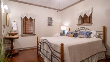 A bedroom with a wooden bed, two windows with white valances, a bedside table with a lamp, and a wall-mounted sign.