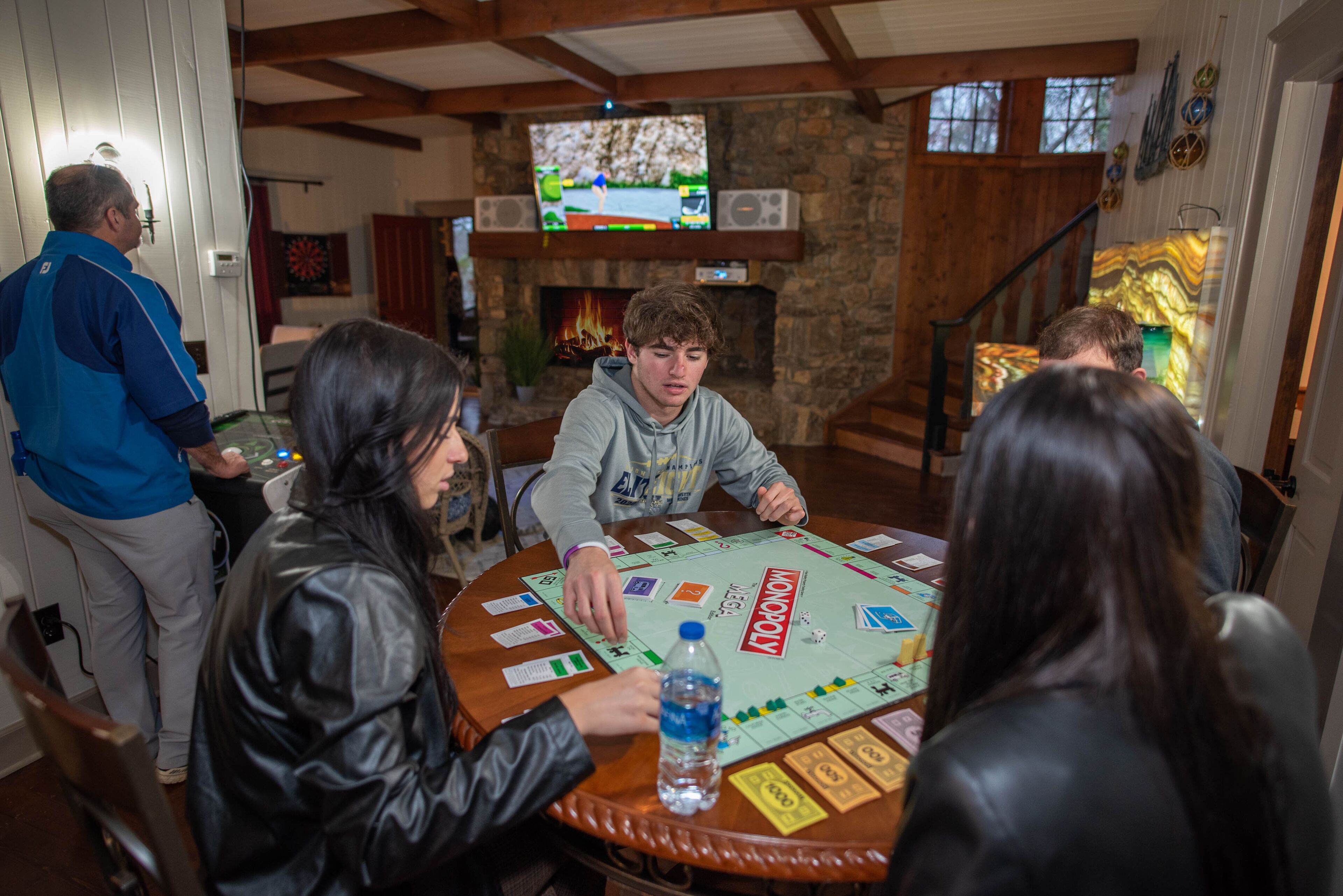 A group of people playing a board game in a room with a stone fireplace, wooden beams, and a television displaying a game.