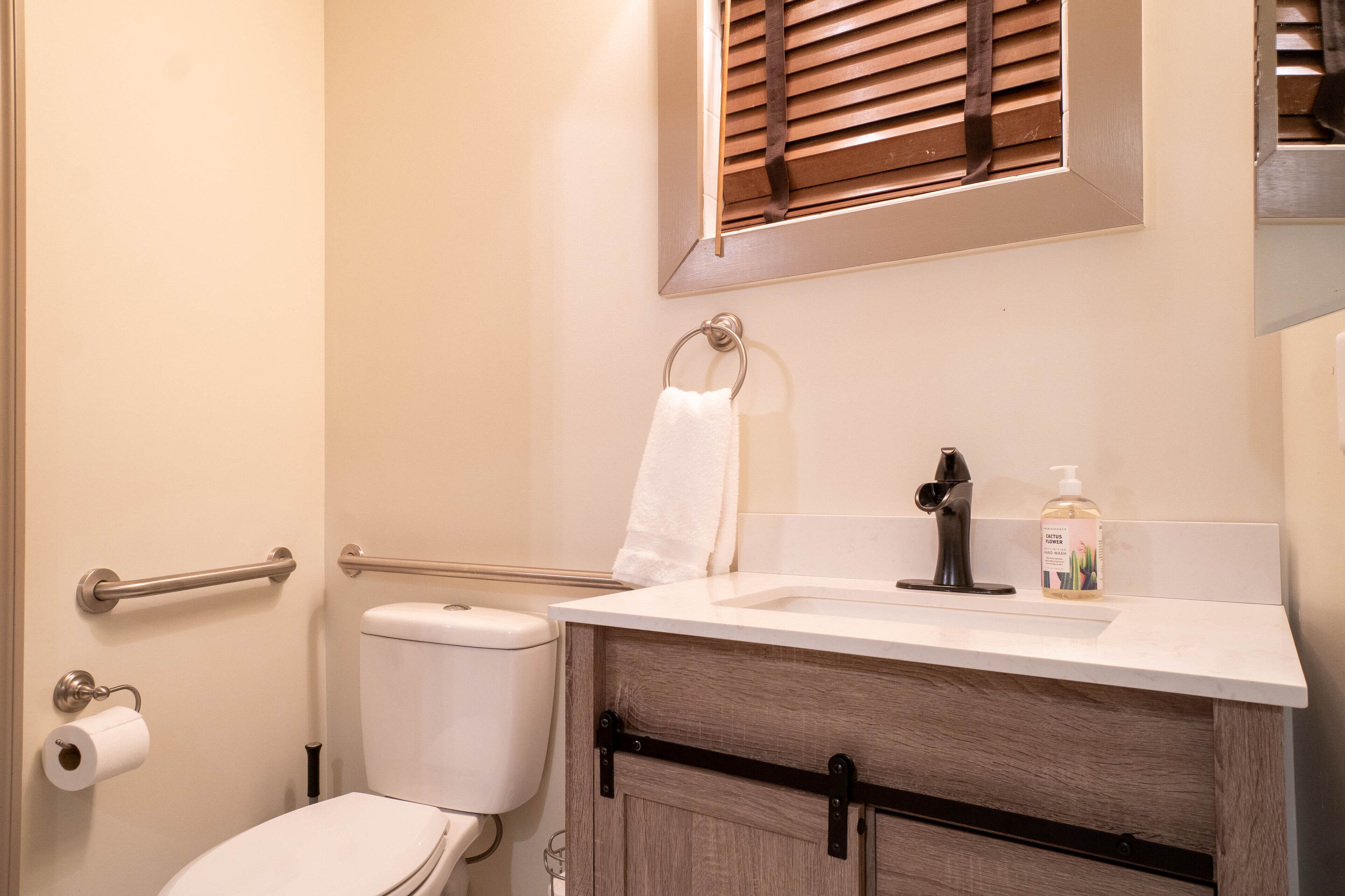 A bathroom with a toilet, a sink with a soap dispenser, a towel hanging on a rack, and a window with blinds.