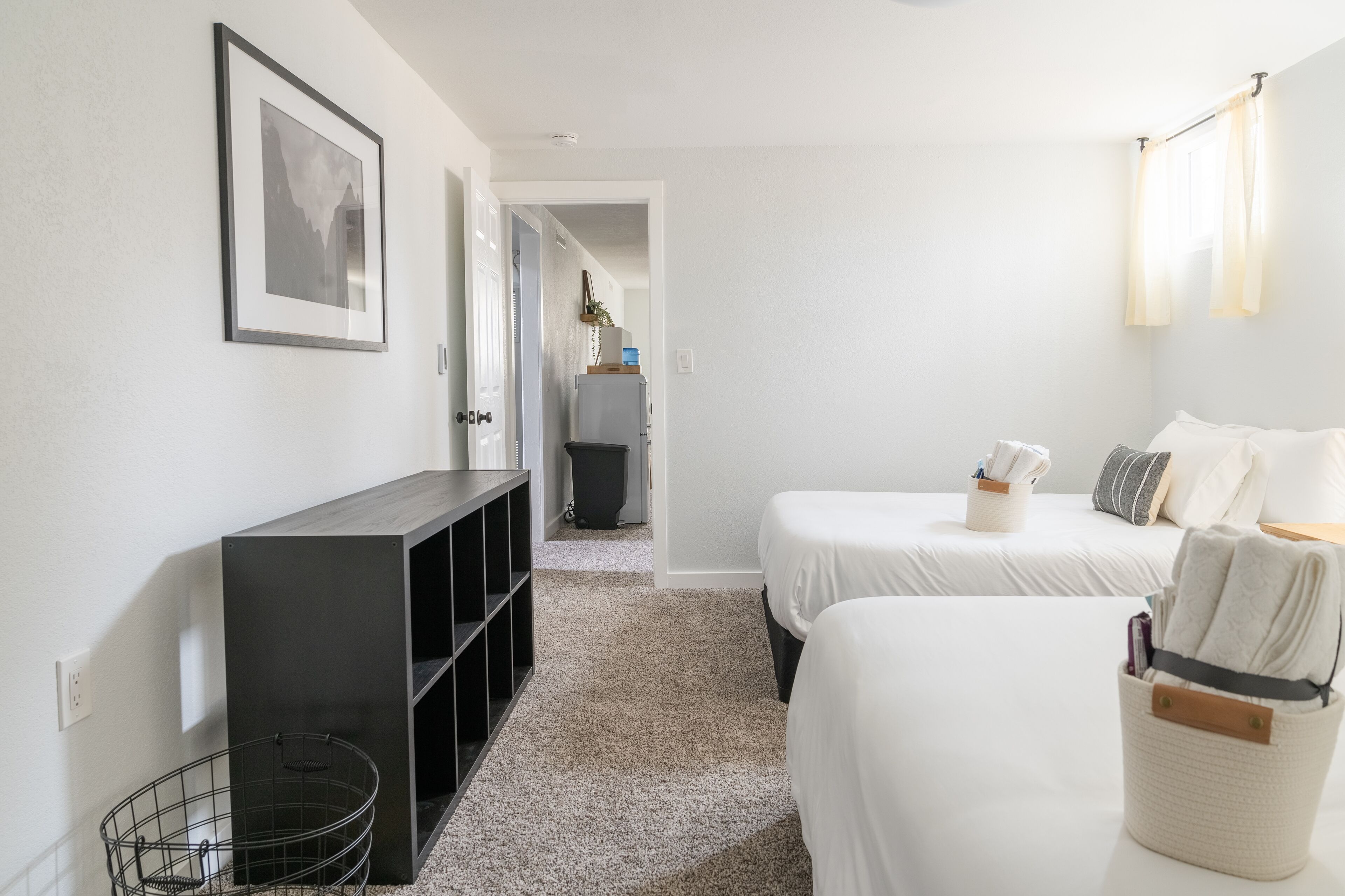 A hotel room with two beds, a small desk, a bookshelf, and a trash bin.