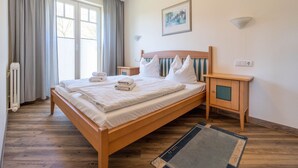A neatly made bed with pillows and folded towels, a wooden nightstand, a radiator, and a window with curtains. - Comfort apartment with 1 bedroom and terrace on the first floor (Ostseebad Binz)