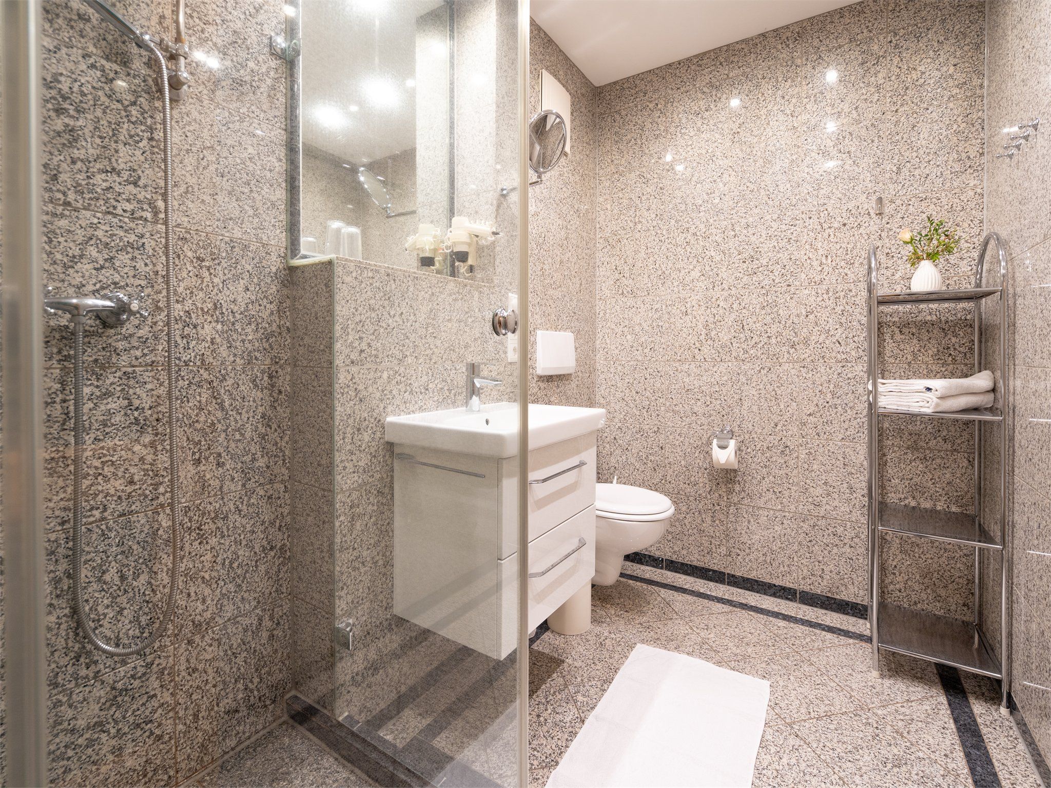 Un baño moderno con ducha envuelta en vidrio, un lavamanos blanco, un inodoro y un perchero con toallas dobladas.