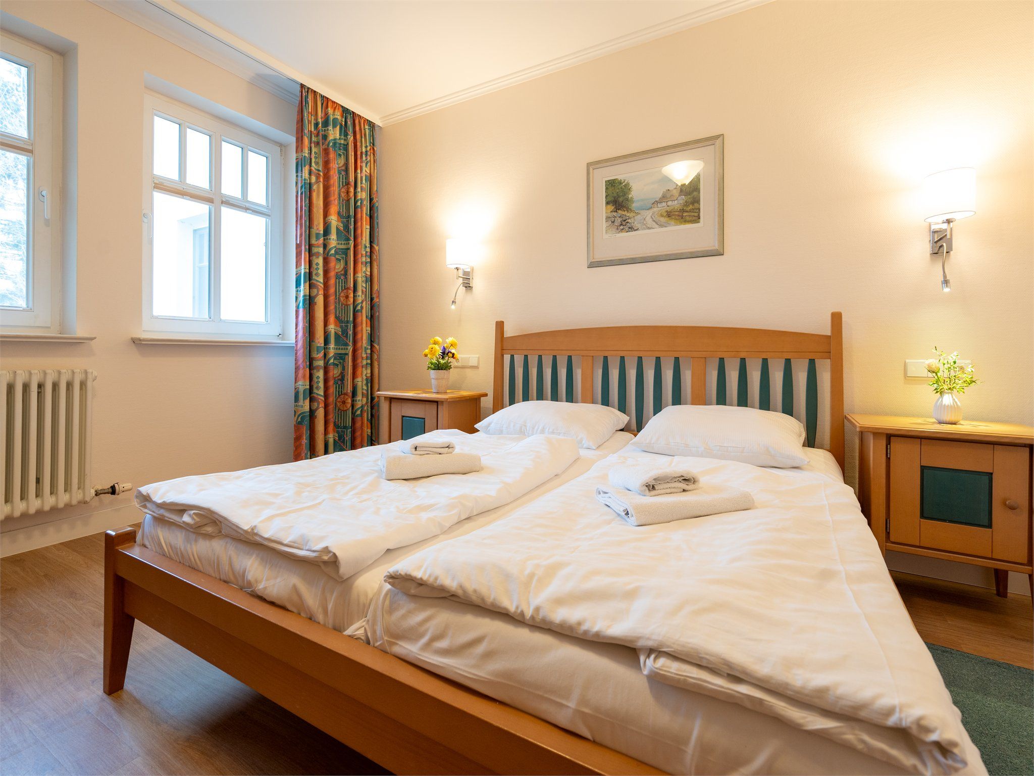 A double bed with white linens, two bedside tables with lamps, a radiator, and a window with curtains.