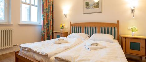 A double bed with white linens, two bedside tables with lamps, a radiator, and a window with curtains.