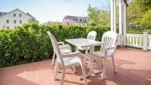 A covered patio with a white table and chairs, surrounded by greenery and a view of houses. - Comfort apartment with 1 bedroom and terrace on the first floor (Ostseebad Binz)