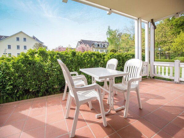 A covered patio with a white table and chairs, surrounded by greenery and a view of houses.
