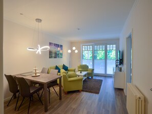 Uma sala de estar moderna com área de jantar, apresentando uma mesa de madeira, cadeiras e um sofá com almofadas. Há grandes janelas com vista para o verde lá fora.