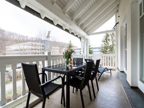 Um alpendre coberto com mesa e cadeiras, um vaso com flores e vista para um edifício e árvores ao fundo.