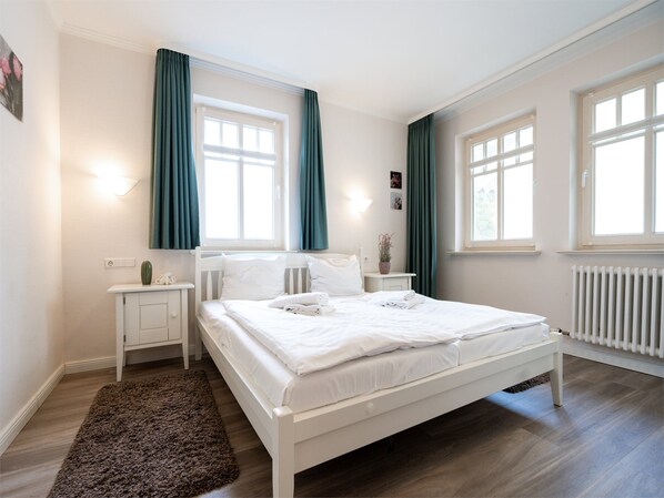 Une chambre avec un lit blanc, deux tables de chevet, une grande fenêtre avec des rideaux, un radiateur et un tapis marron.