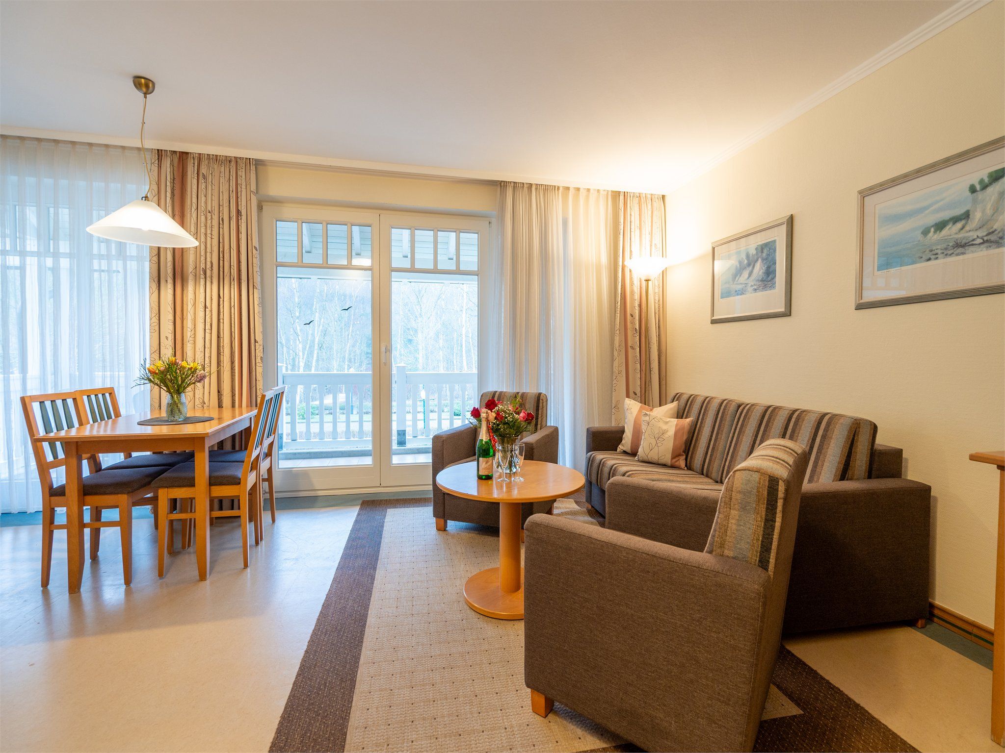 Un salon comprenant un canapé, des fauteuils, une table à manger avec des chaises, une table basse et une grande fenêtre avec des rideaux.