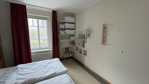 A hotel room with a bed, a radiator, a window with curtains, a chair, a shelf with boxes, a mirror, and a wall-mounted light fixture. - Comfort apartment with 1 bedroom and terrace on the first floor (Ostseebad Binz)