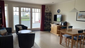 A living room with a sofa, armchair, and a dining table with chairs. There is a television on a stand and a bookshelf filled with books. - Comfort apartment with 1 bedroom and terrace on the first floor (Ostseebad Binz)