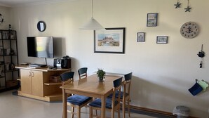A dining area with a wooden table and chairs, a television on a cabinet, and wall decorations including framed pictures and a clock. - Comfort apartment with 1 bedroom and terrace on the first floor (Ostseebad Binz)