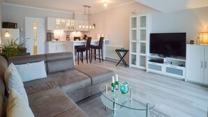 A modern living room with a sofa, a glass coffee table, and a TV. There is a kitchen area with cabinets and a dining table. - Comfort plus apartment with 1 bedroom and balcony on the top floor (Ostseebad Binz)