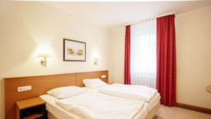 A hotel room with two beds, a nightstand, a lamp, a framed picture on the wall, and a window with red curtains. - Komfort Ferienwohnung mit 1 Schlafzimmer und Balkon im Obergeschoss (Ostseebad Binz)