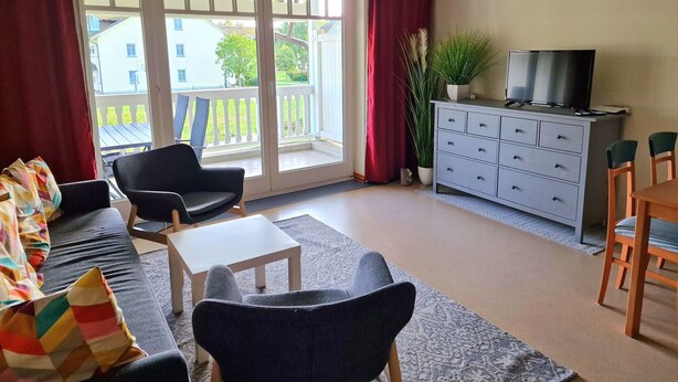 A living room with a sofa, chairs, a TV, a dining table, and a balcony with a view of buildings and trees. - Komfort Ferienwohnung mit 1 Schlafzimmer und Balkon im Obergeschoss (Ostseebad Binz)