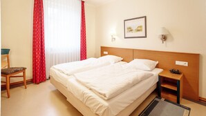 A hotel room with a double bed, a chair, a bedside table with a lamp, and a window with red curtains. - Comfort apartment with 1 bedroom and balcony on the upper floor (Ostseebad Binz)