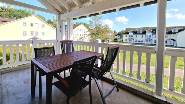A covered outdoor area with a table and chairs, a white railing, and a view of residential buildings. - Comfort apartment with 1 bedroom and balcony on the upper floor (Ostseebad Binz)