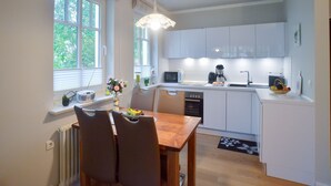 A modern kitchen with white cabinets, a wooden dining table, and a window with blinds. - Comfortplus vacation apartment with 1 bedroom and terrace on the first floor (Ostseebad Binz)